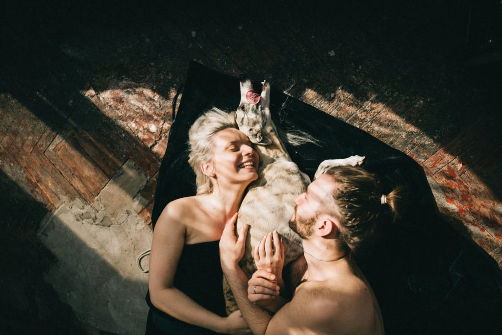 dog cuddling with man and woman, lying on floor, bare chested, titled floor, black blanket, sun shining // Healthier Pets Today