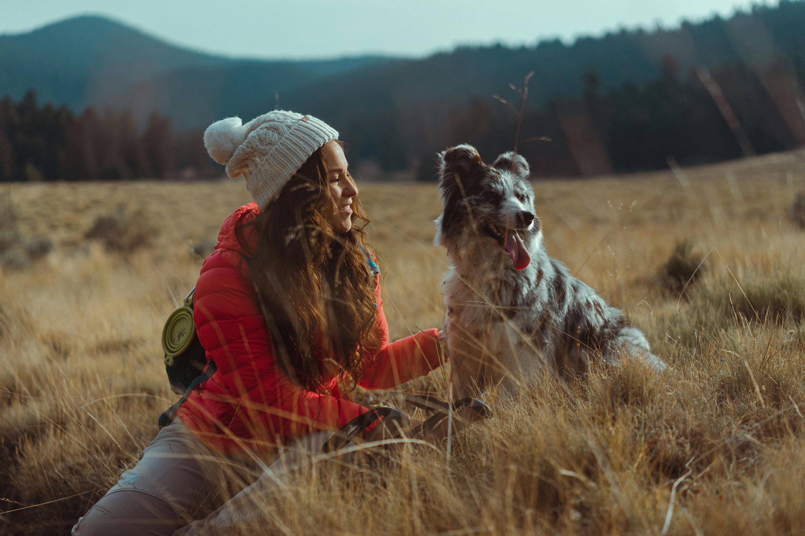 A Woman Sitting with Her Dog in a field, wearing red jacket, white beanie, mountains at the back // Healthier Pets Today