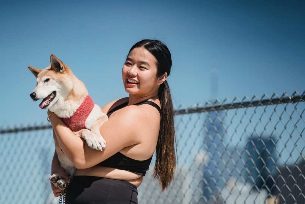woman wit dark long hair, black wokrut clothes, holding fluffy dog, fence behind, clear blue sky // Healthier Pets Today