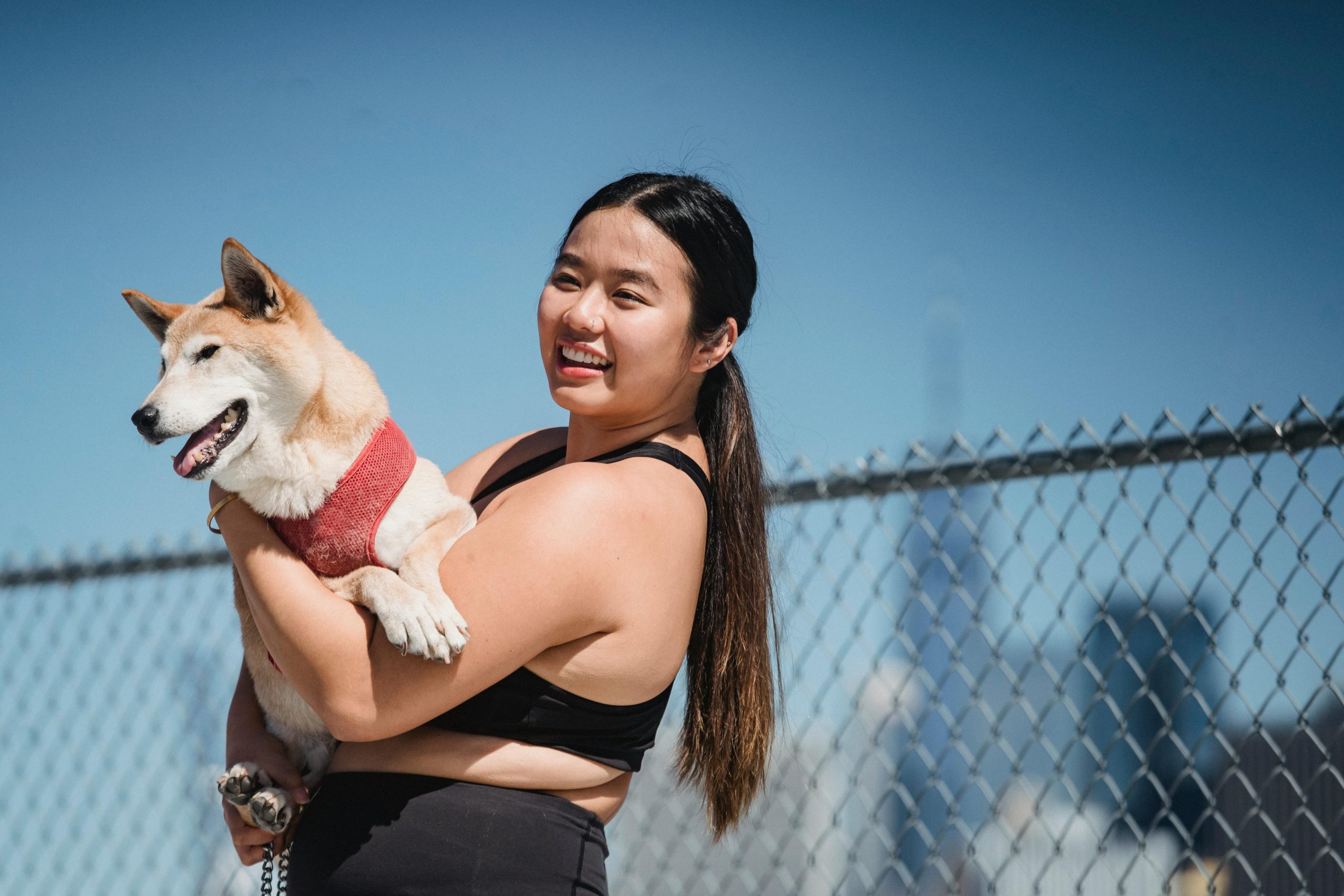 woman wit dark long hair, black wokrut clothes, holding fluffy dog, fence behind, clear blue sky // Healthier Pets Today